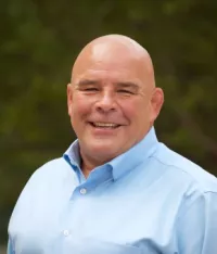 Bald man smiling wearing a light blue shirt against a natural background