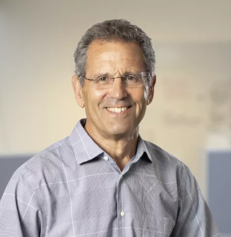 Older man wearing a grey buttoned shirt and glasses with a smile against an industrial background