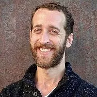 smiling man with a beard wearing a black sweater against an industrial backgound